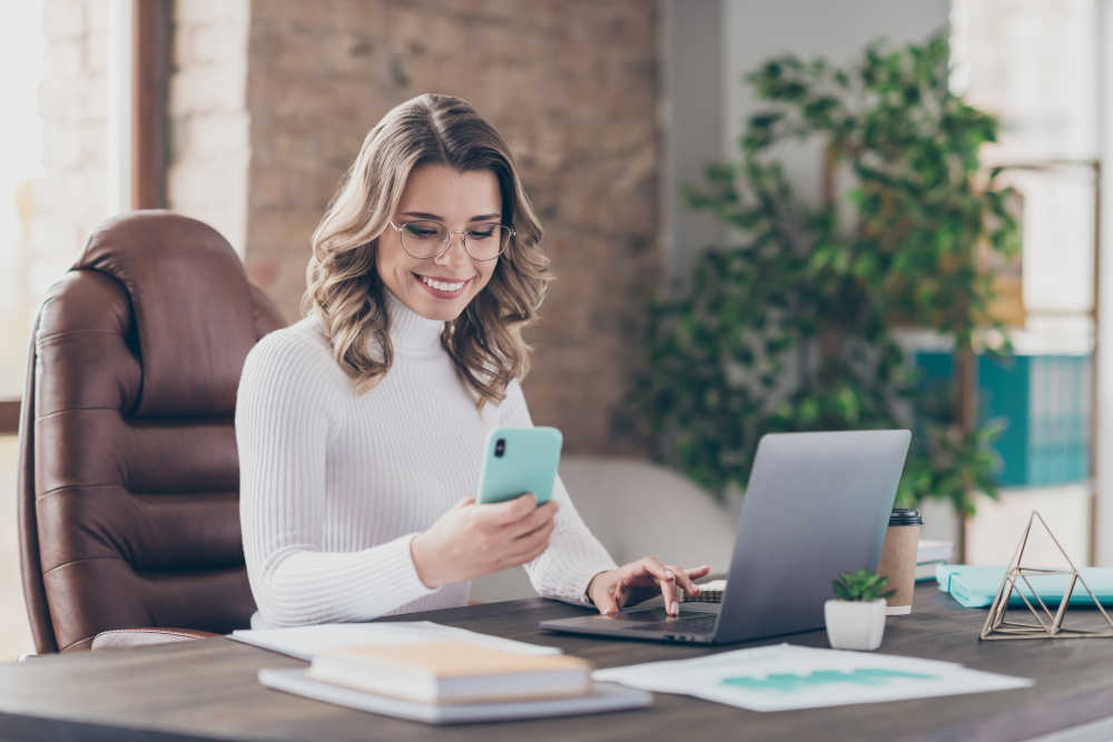 A smiling woman wearing glasses and a white turtleneck sweater sits at a desk in a stylish office, multitasking between her smartphone and laptop. The workspace is well-organized with documents, a coffee cup, a small potted plant, and decorative elements, creating a productive and modern work environment.